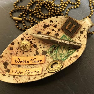 Op de afbeelding: Een vintage lepelketting met een bruine leren initiaalbedel, een gouden penpunt en een label met de tekst "Schrijf je eigen verhaal".