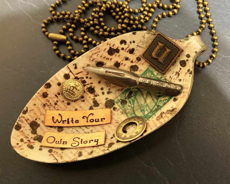Op de afbeelding: Een vintage lepelketting met een bruine leren initiaalbedel, een gouden penpunt en een label met de tekst "Schrijf je eigen verhaal".