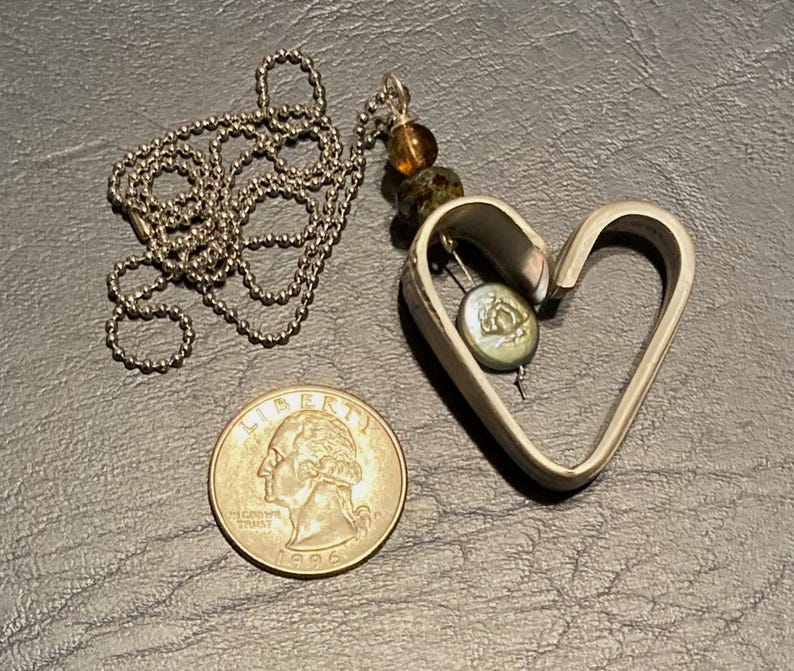 May include: A silver heart-shaped pendant necklace with a silver chain. The pendant has a small green and brown bead and a silver charm inside.