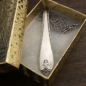 Puede incluir: Un collar con un colgante de cuchara de plata con un dise&ntilde;o floral en la parte inferior. El collar est&aacute; en una peque&ntilde;a caja de regalo dorada.