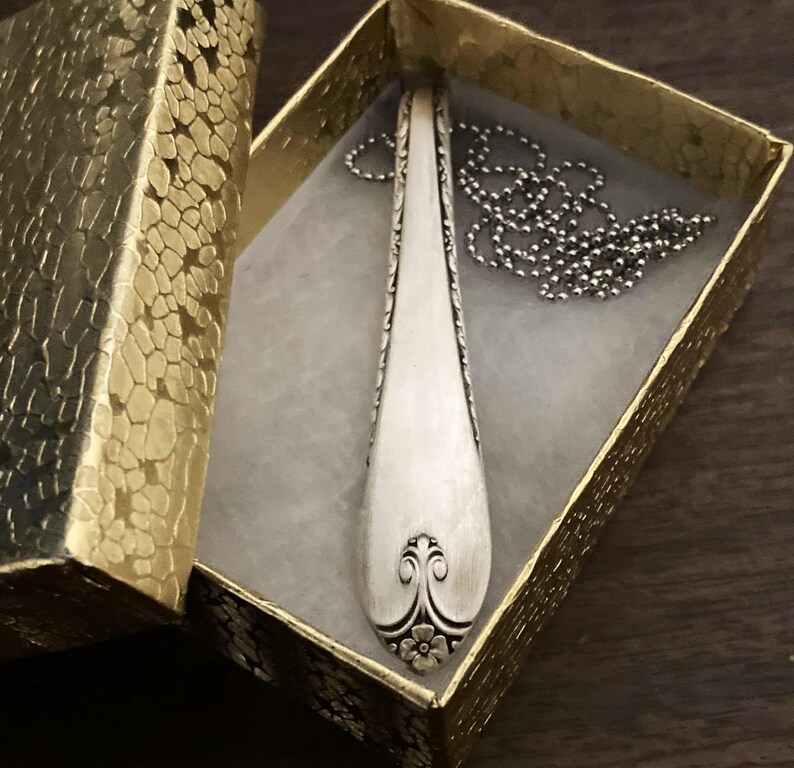 Puede incluir: Un collar con un colgante de cuchara de plata con un dise&ntilde;o floral en la parte inferior. El collar est&aacute; en una peque&ntilde;a caja de regalo dorada.