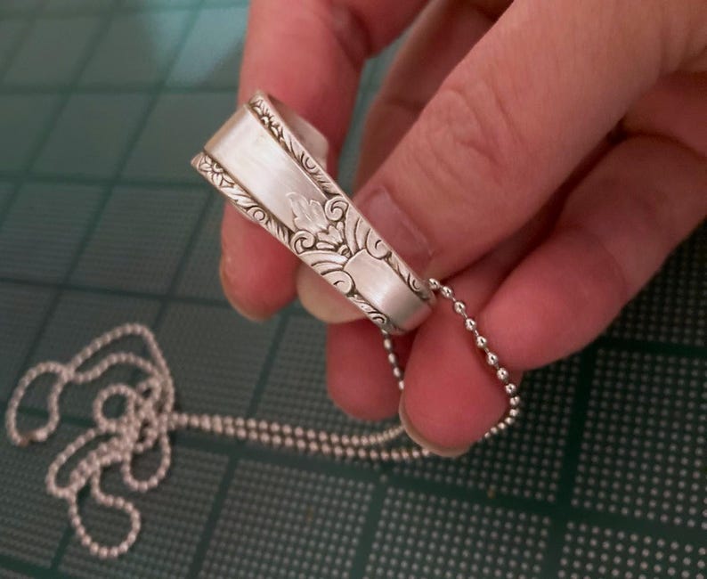 Puede incluir: Un collar de plata con una cuchara adornada con un dise&ntilde;o floral detallado. El collar est&aacute; hecho con una cadena de bolas de plata.