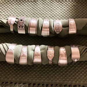 May include: A set of 14 silver napkin rings made from repurposed silverware. The rings are arranged in two rows on a green fabric background.