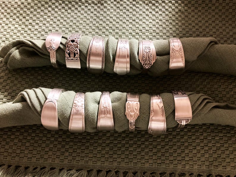May include: A set of 14 silver napkin rings made from repurposed silverware. The rings are arranged in two rows on a green fabric background.