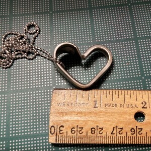 May include: A silver heart-shaped pendant necklace with a ball chain. The pendant is open on one side, creating a heart shape. The necklace is measured against a ruler, showing it is approximately 1 inch in length.