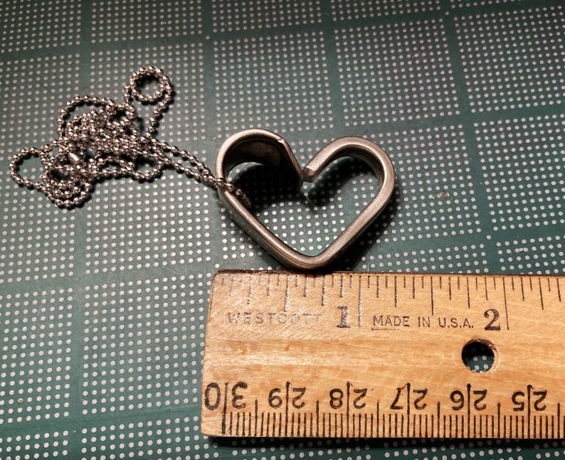 May include: A silver heart-shaped pendant necklace with a ball chain. The pendant is open on one side, creating a heart shape. The necklace is measured against a ruler, showing it is approximately 1 inch in length.