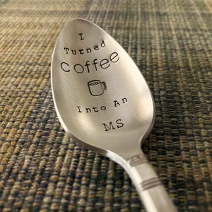Pu&ograve; includere: Un cucchiaio in acciaio inossidabile con la scritta "I Turned Coffee Into An MS" incisa sulla conca del cucchiaio. Una piccola grafica di tazza di caff&egrave; &egrave; tra le parole "Coffee" e "Into". Il cucchiaio &egrave; su una superficie strutturata.