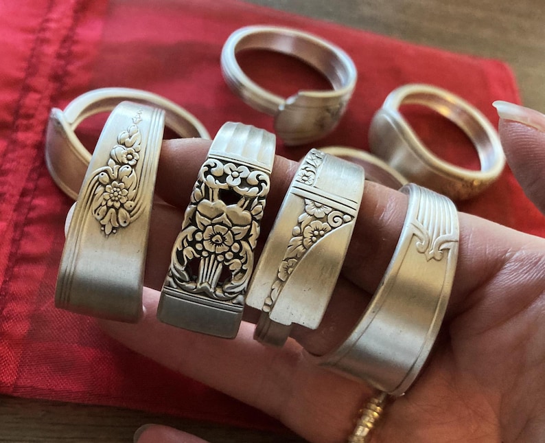 May include: Six silver napkin rings with ornate designs. The rings are stacked on top of each other and are being held by a hand.