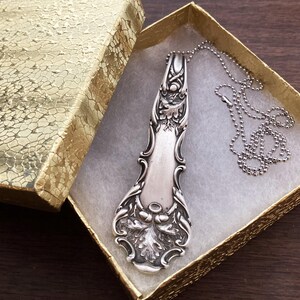 May include: A silver vintage spoon necklace with an ornate design, hanging in a gold gift box with white lining.