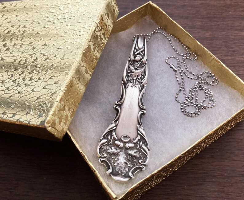 May include: A silver vintage spoon necklace with an ornate design, hanging in a gold gift box with white lining.