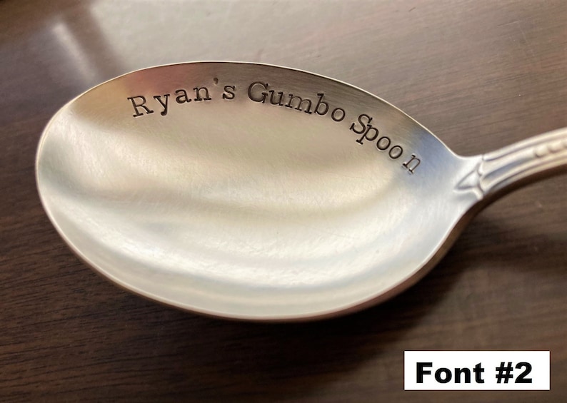May include: A silver spoon with the text "Ryan's Gumbo Spoon" engraved on the bowl. The spoon is on a wooden surface. Font #2.