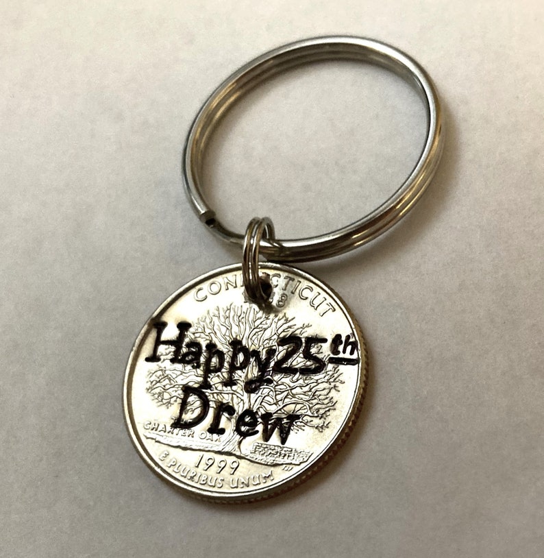 May include: A silver keychain with a quarter coin pendant. The coin is engraved with "Happy 25th Drew" and a tree design. The coin also has the text "Connecticut" and "1999".