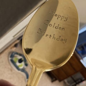 May include: A gold-colored spoon with the words "Happy Golden Birthday" engraved on the spoon's bowl. The spoon has a shiny, reflective surface and a long handle. The text is in a simple, sans-serif font.