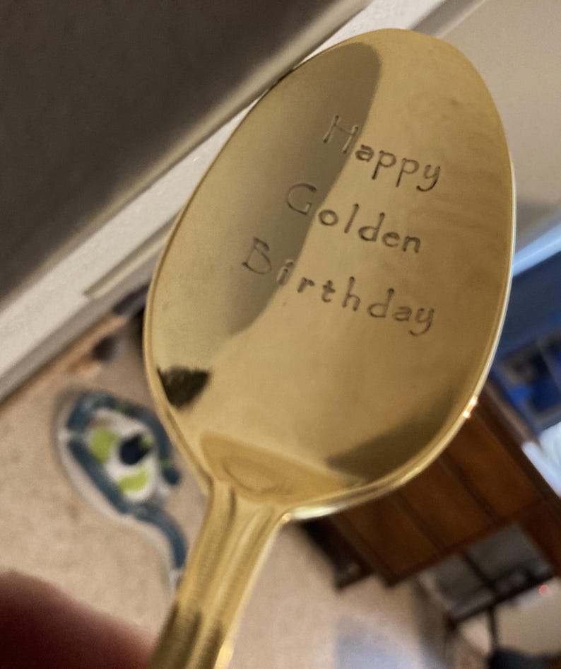 May include: A gold-colored spoon with the words "Happy Golden Birthday" engraved on the spoon's bowl. The spoon has a shiny, reflective surface and a long handle. The text is in a simple, sans-serif font.