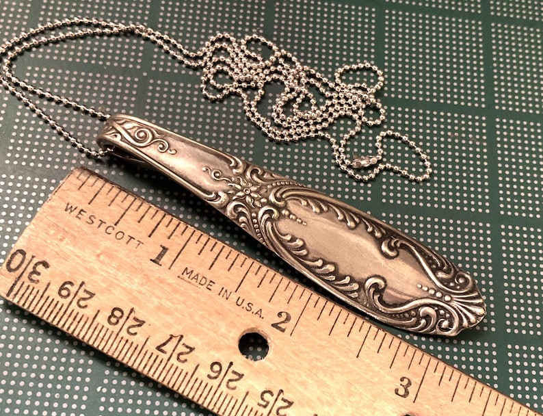 May include: A silver spoon necklace with an ornate design. The spoon is about 2 inches long and is attached to a silver chain. The chain is about 18 inches long and has a lobster clasp.