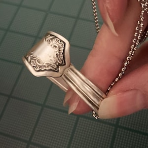 May include: A silver spoon ring with an ornate design. The ring is attached to a silver chain.