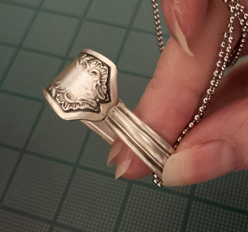 May include: A silver spoon ring with an ornate design. The ring is attached to a silver chain.