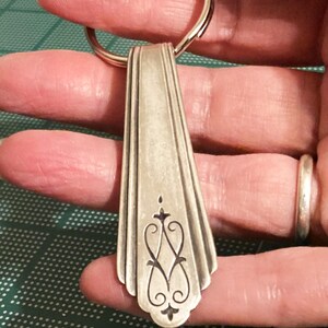 May include: A silver spoon keychain with an ornate design on the handle. The spoon is attached to a silver key ring.