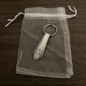 May include: A silver spoon keychain with an ornate design, packaged in a white organza bag.