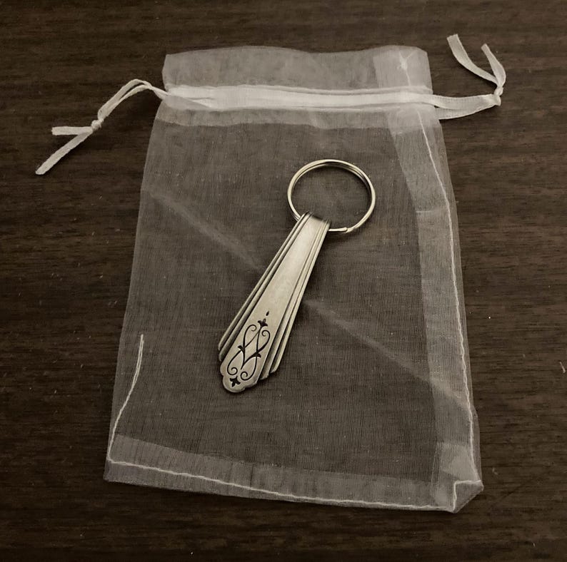 May include: A silver spoon keychain with an ornate design, packaged in a white organza bag.