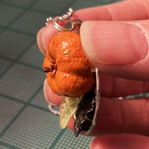 May include: A silver chain necklace with a pendant featuring a small orange pumpkin, a black crow, and a green leaf.