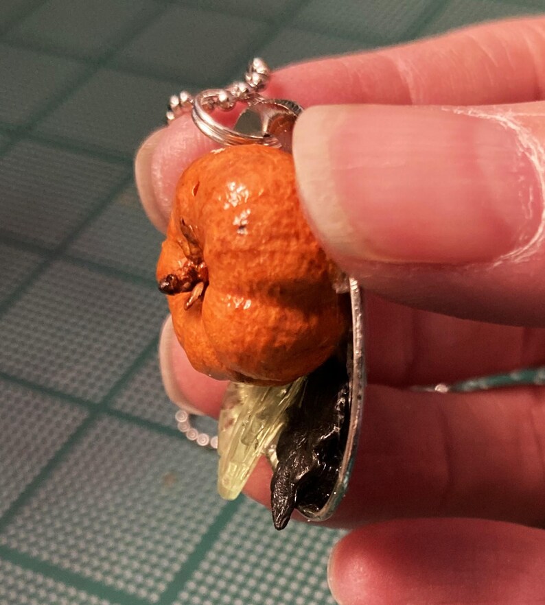 May include: A silver chain necklace with a pendant featuring a small orange pumpkin, a black crow, and a green leaf.