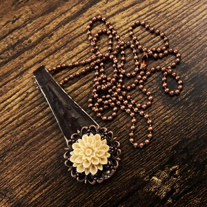 May include: A copper chain necklace with a pendant featuring a brown, textured, rectangular piece with a cream-coloured flower.