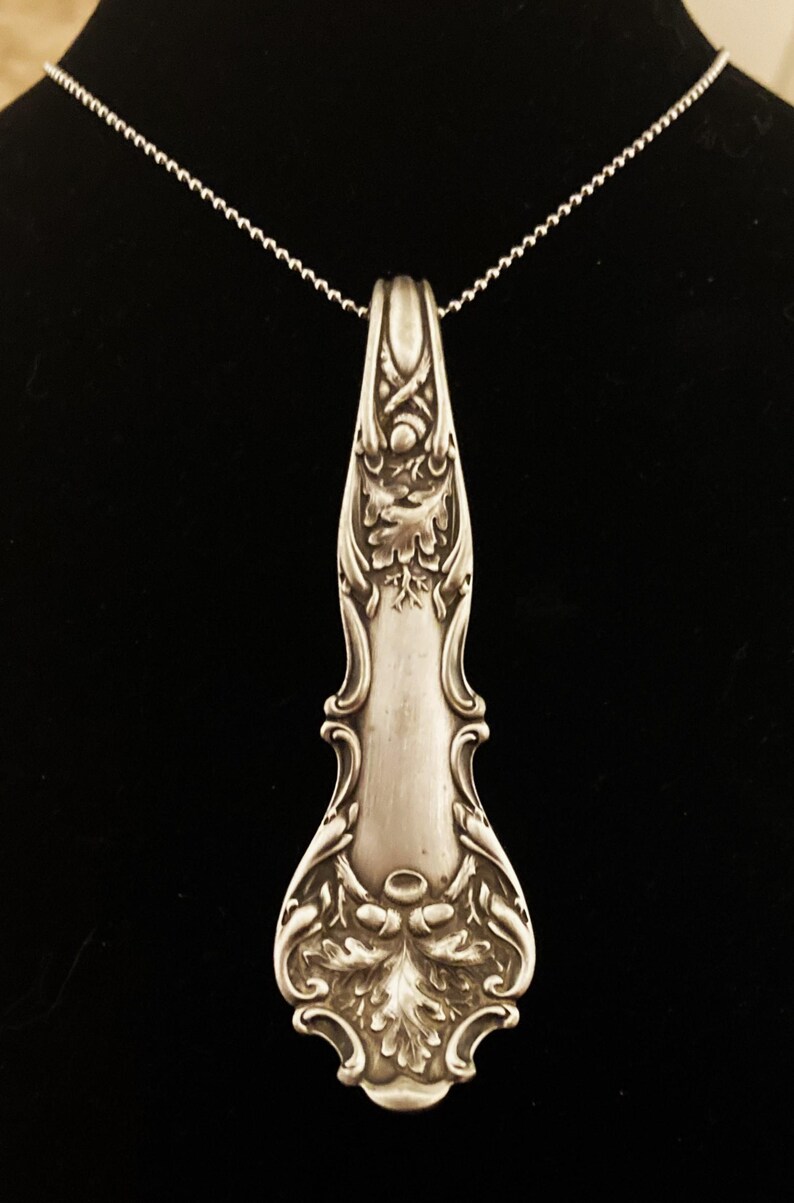 May include: A silver necklace with a pendant made from an ornate antique spoon. The spoon has an intricate design of leaves and swirls.