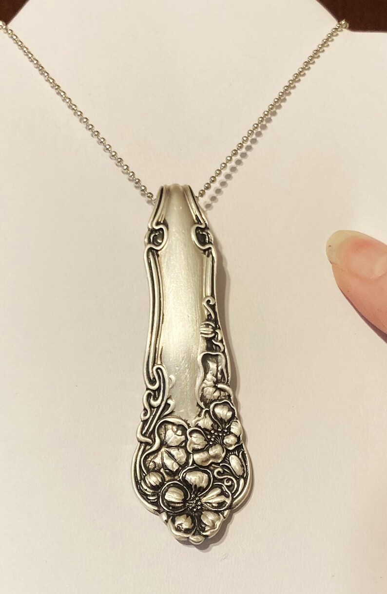 May include: A silver necklace with a pendant made from a vintage spoon. The spoon has a floral design and is polished to a shine.