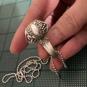 May include: A silver spoon ring with a grape design, attached to a silver ball chain necklace.