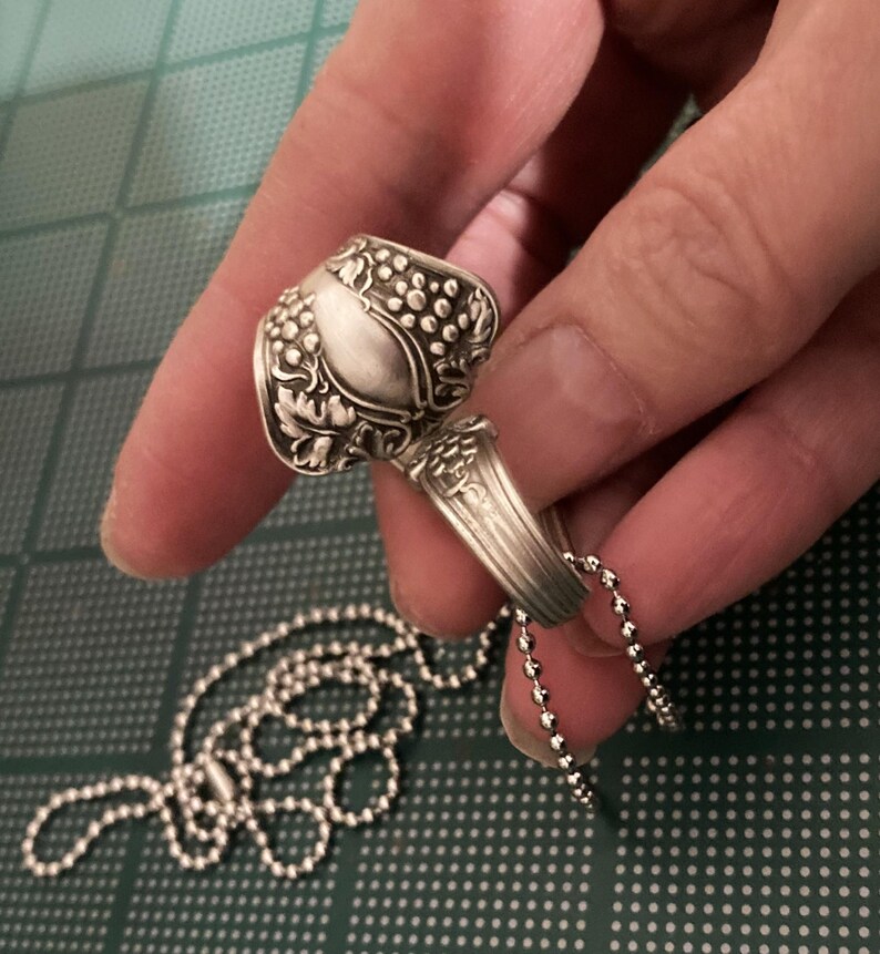 May include: A silver spoon ring with a grape design, attached to a silver ball chain necklace.