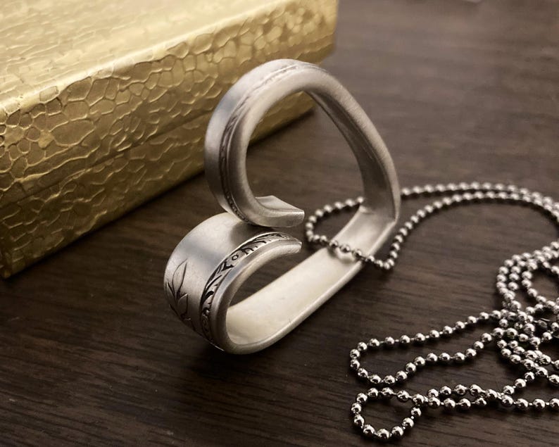 May include: A silver heart-shaped pendant necklace made from a repurposed spoon. The pendant has a detailed floral design and is attached to a silver ball chain.