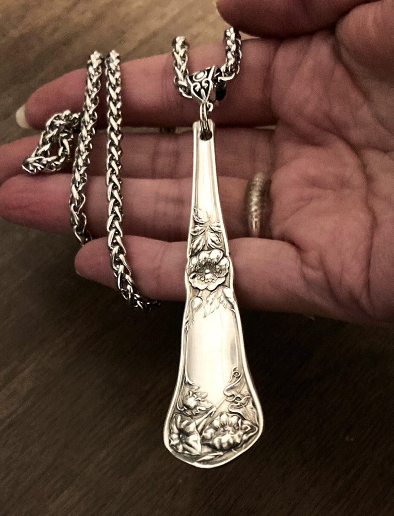 May include: A silver necklace with a long, ornate pendant made from a vintage silverware spoon. The pendant features a floral design with a large flower in the center.