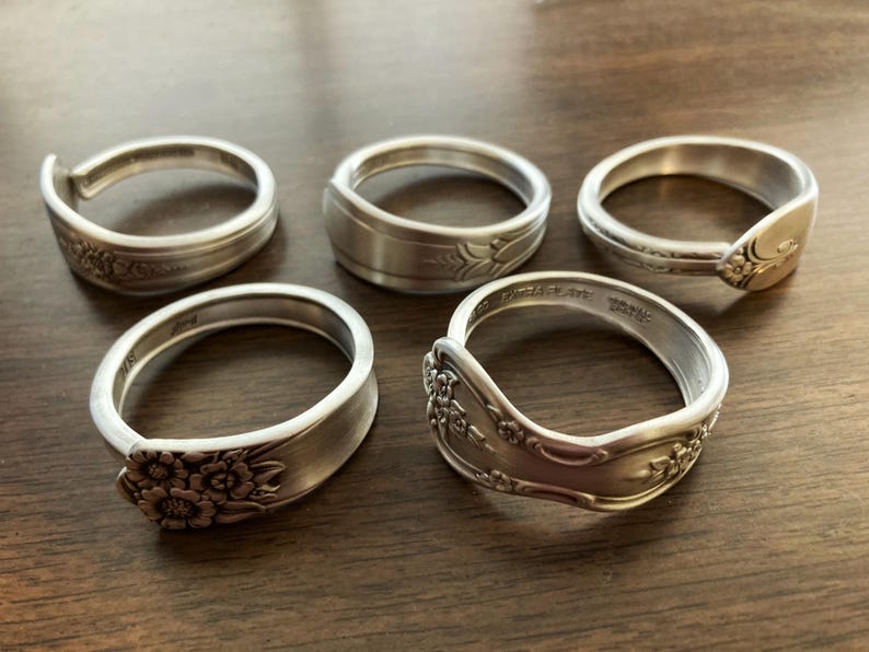 Puede incluir: Cinco anillos de servilleta de plata hechos con cubiertos reutilizados con dise&ntilde;os florales ornamentados.