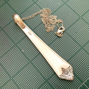 May include: A silver pendant necklace crafted from a repurposed spoon. The pendant features a floral design at the base and is attached to a delicate chain. The spoon's handle has a small floral accent. The necklace rests on a green surface.