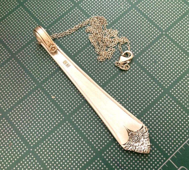 May include: A silver pendant necklace crafted from a repurposed spoon. The pendant features a floral design at the base and is attached to a delicate chain. The spoon's handle has a small floral accent. The necklace rests on a green surface.