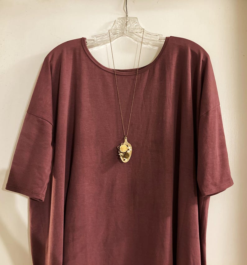 May include: A maroon colored, loose-fitting, short-sleeved t-shirt with a gold spoon pendant necklace. The spoon has a floral design.
