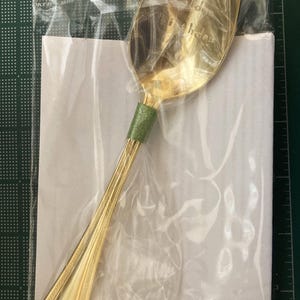 May include: A gold-colored spoon with a green handle wrap, packaged in a clear plastic bag. The spoon has a decorative design on the handle and the words "Happy Birthday" are visible on the spoon's bowl.