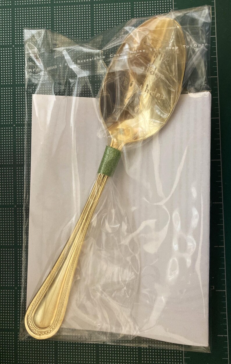 May include: A gold-colored spoon with a green handle wrap, packaged in a clear plastic bag. The spoon has a decorative design on the handle and the words "Happy Birthday" are visible on the spoon's bowl.
