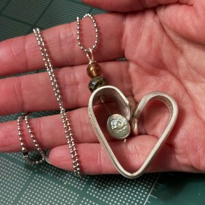 May include: A silver heart-shaped pendant necklace with a smaller heart charm inside. The necklace is on a silver ball chain.