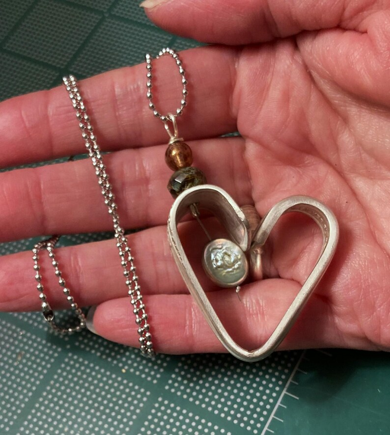 May include: A silver heart-shaped pendant necklace with a smaller heart charm inside. The necklace is on a silver ball chain.