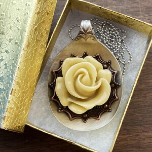 May include: A silver spoon pendant necklace with a white rose made of polymer clay. The rose is surrounded by a copper-colored filigree design. The necklace is displayed in a gold gift box.