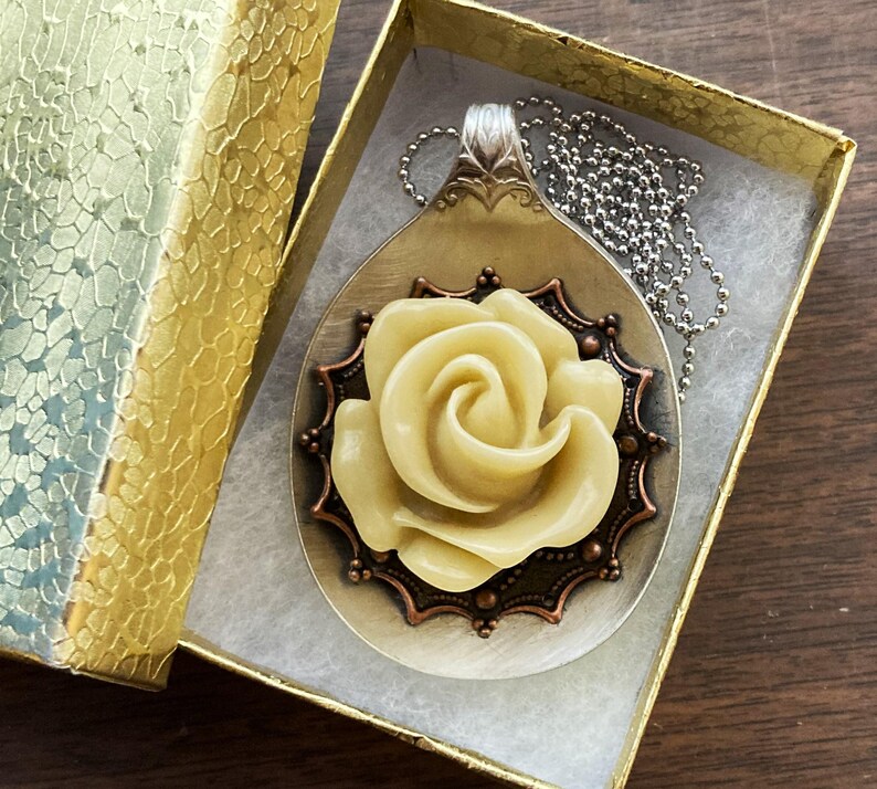 May include: A silver spoon pendant necklace with a white rose made of polymer clay. The rose is surrounded by a copper-colored filigree design. The necklace is displayed in a gold gift box.