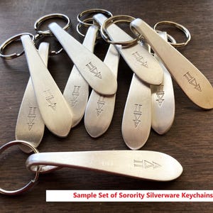 May include: A group of silver metal keyrings, each with a spoon-shaped design and a ring attachment. The keyrings have an engraved symbol and the text "Sample Set of Sorority Silverware Keychains" is visible.