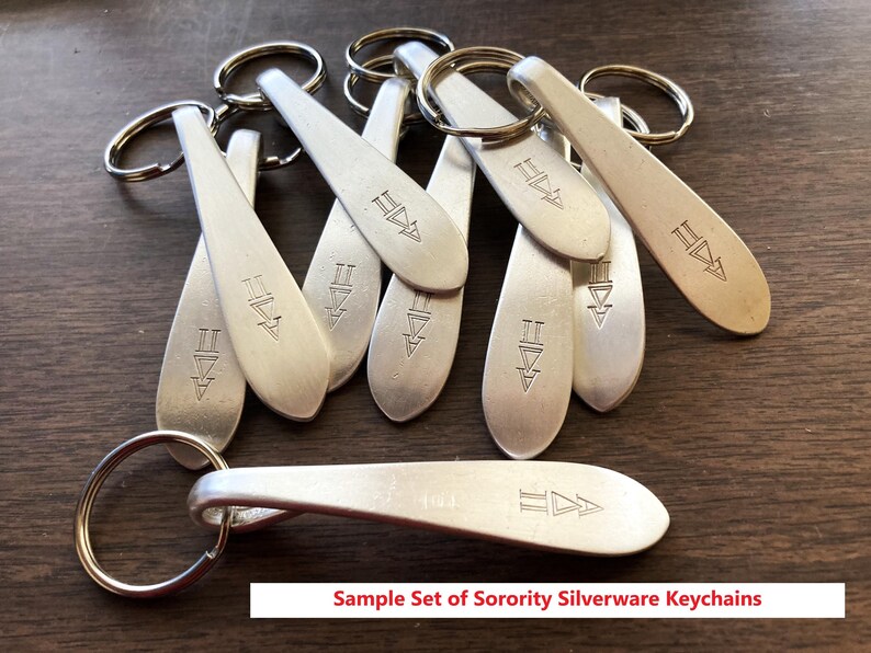 May include: A group of silver metal keyrings, each with a spoon-shaped design and a ring attachment. The keyrings have an engraved symbol and the text "Sample Set of Sorority Silverware Keychains" is visible.