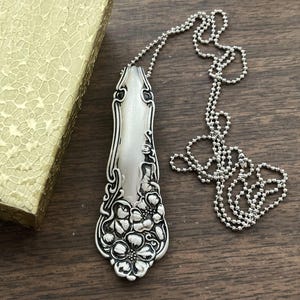 May include: A silver spoon necklace with a floral design. The spoon is attached to a silver ball chain.