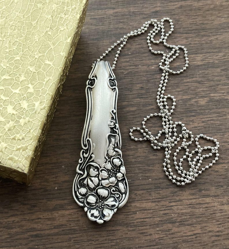 May include: A silver spoon necklace with a floral design. The spoon is attached to a silver ball chain.
