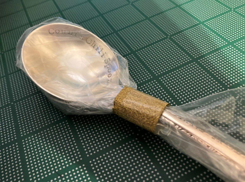 May include: A silver chili spoon with the words "Come get a Chili Spoon" engraved on the bowl. The spoon is wrapped in clear plastic and a brown tape.
