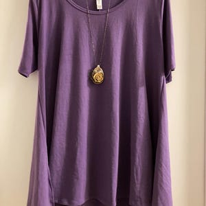 May include: A purple, loose-fitting, short-sleeved t-shirt with a longer back hem. The shirt is hanging on a white hanger and has a silver chain necklace with a rose-shaped pendant.