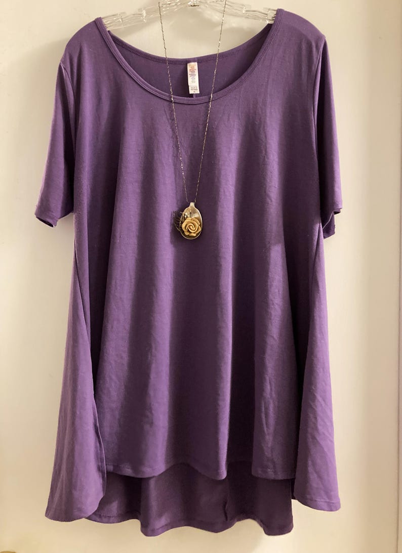 May include: A purple, loose-fitting, short-sleeved t-shirt with a longer back hem. The shirt is hanging on a white hanger and has a silver chain necklace with a rose-shaped pendant.